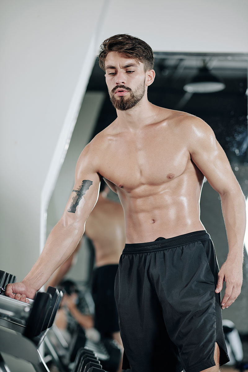 A lean shirtless man with tidy facial hair reaches for a dumbbell