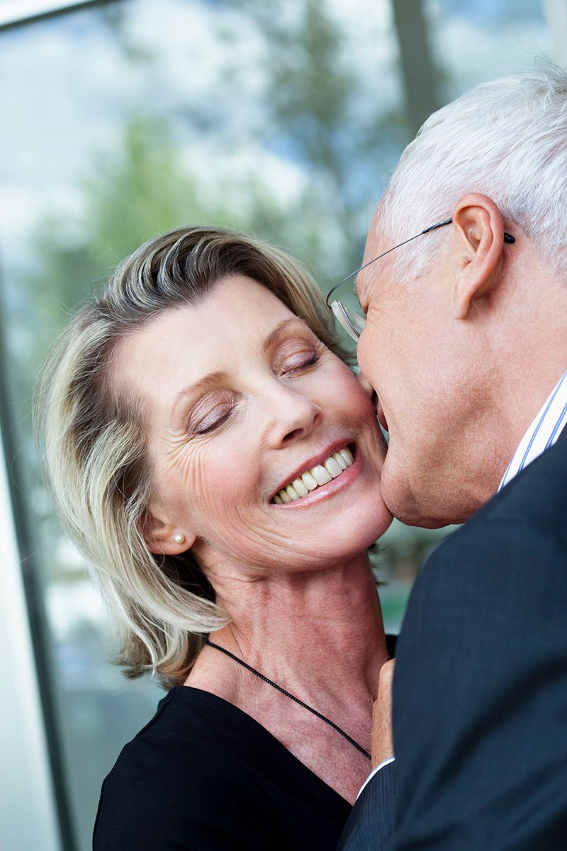 A man with white hair and glasses leans in to kiss or whisper to his smiling wife