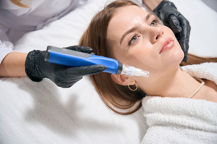 A doctor with gloved hands performs microneedling services on a young woman’s face using a SkinPen