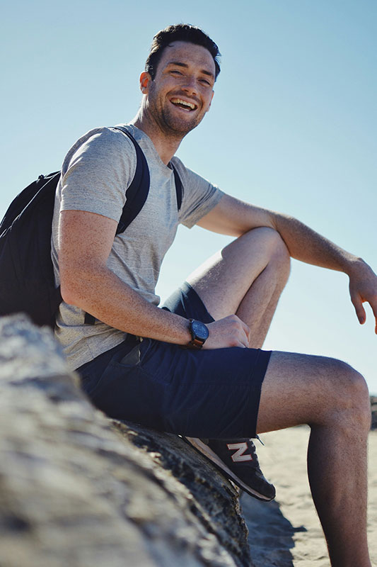 A smiling hiker wearing athletic clothing and a backpack sits against a rock