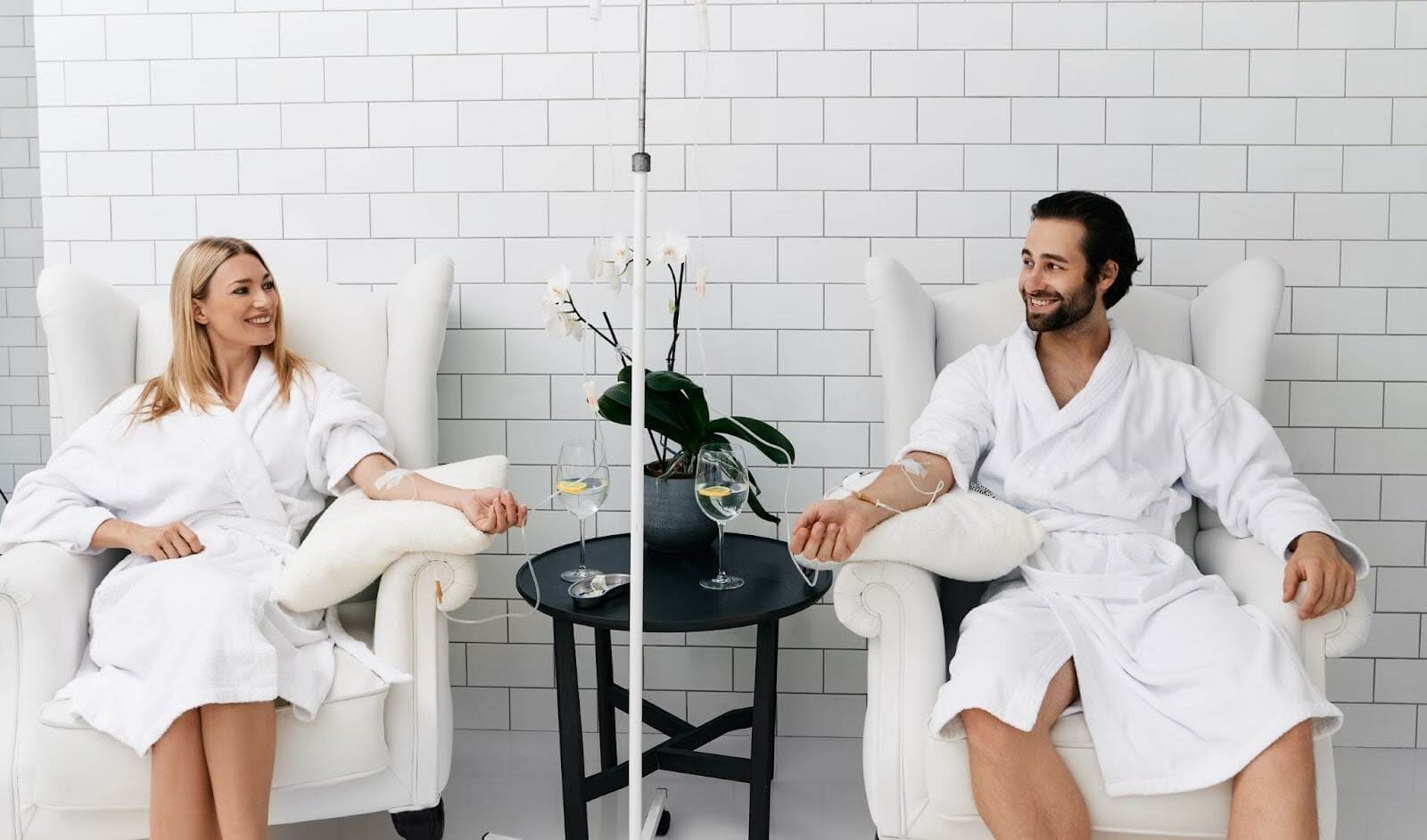 A smiling couple in matching white robes exchanges glances while they sit and receive vitamin IV therapy