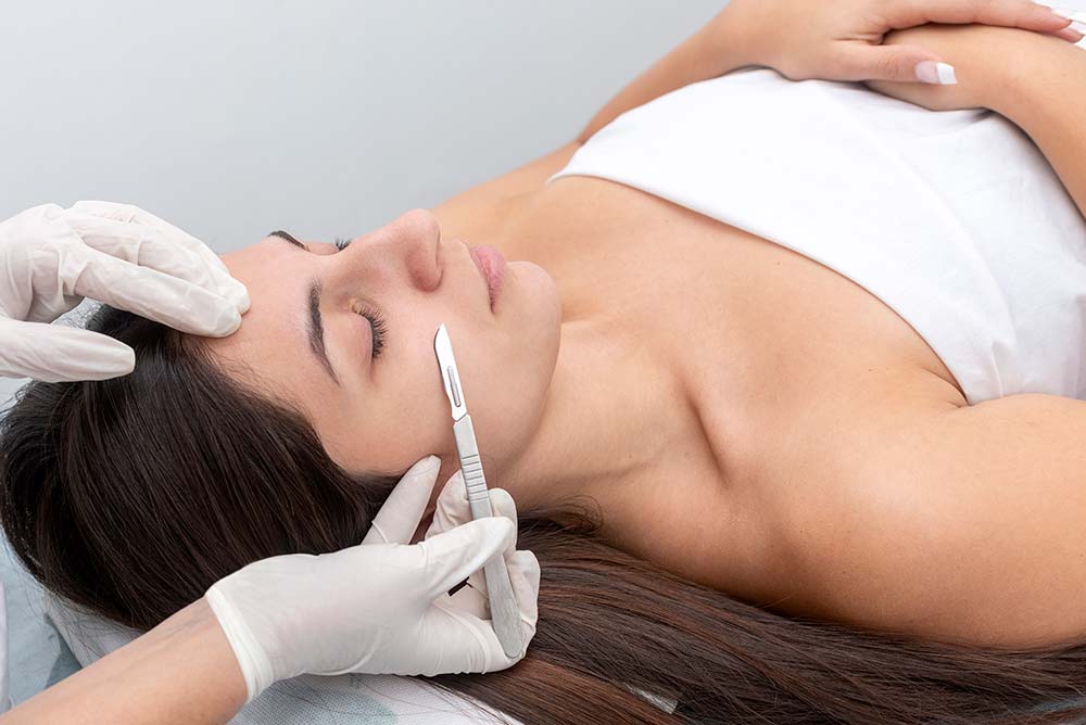 A scalpel is held against the cheek of a med spa patient during a dermaplaning treatment
